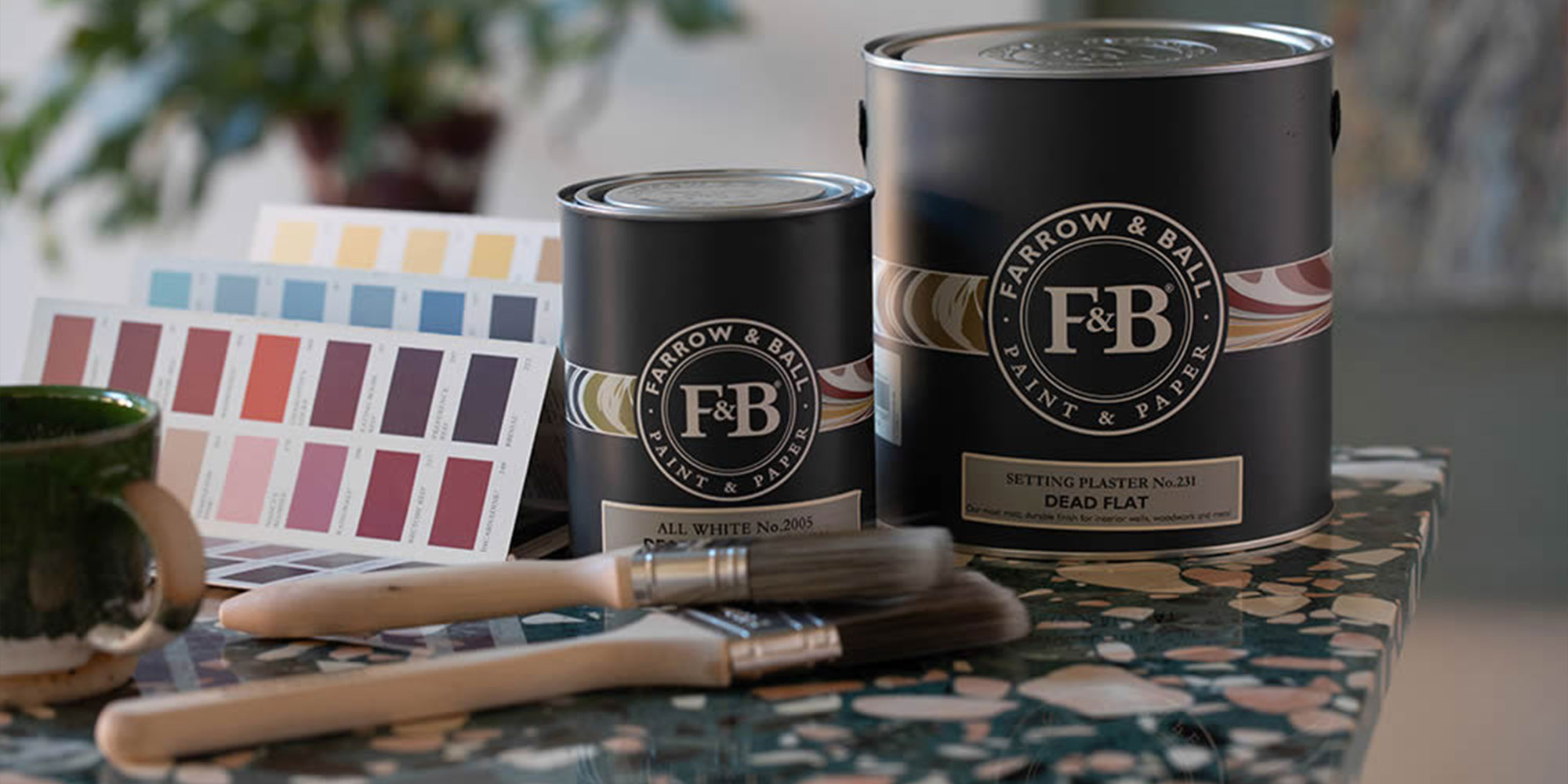 Zwei Farbdosen von Farrow and Ball mit Farbkarte und Pinseln neben Tasse auf gemusterter Tischplatte