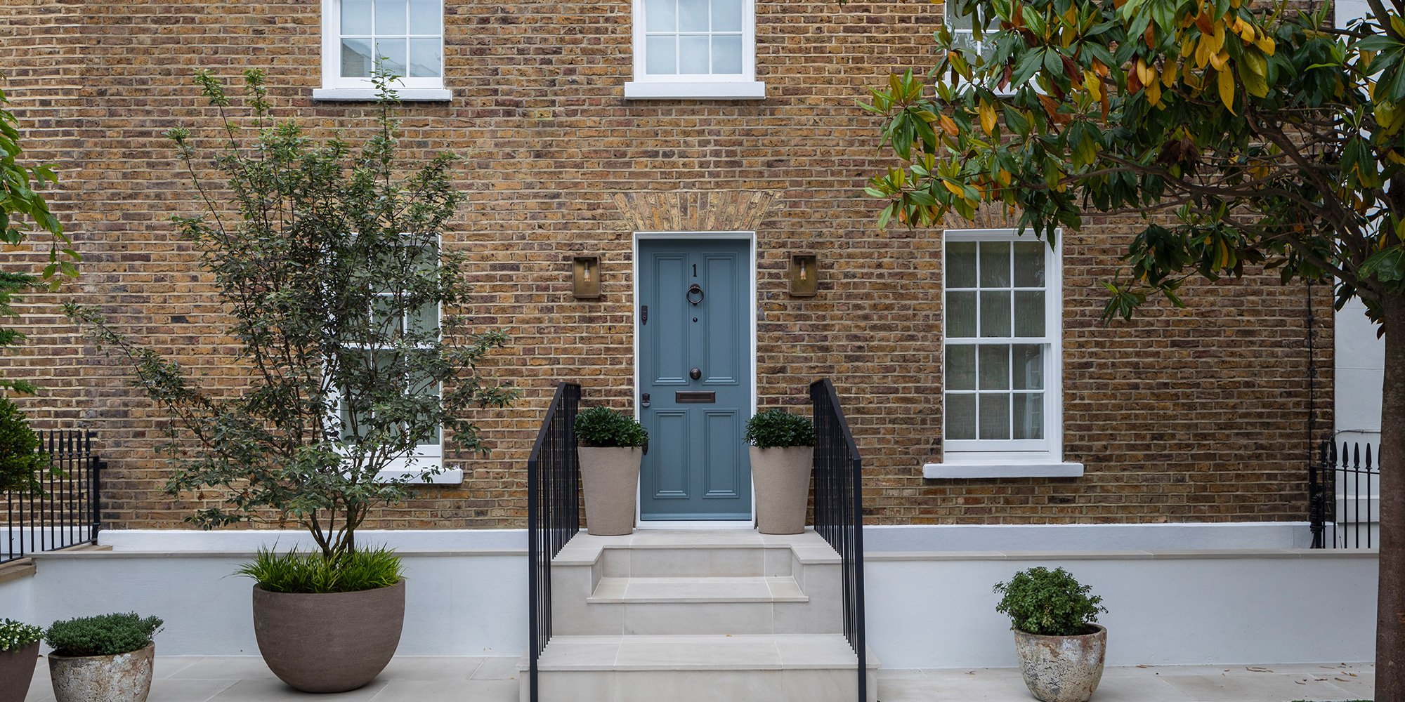 Blaue Haustür an Backsteinfassade mit Treppe, Geländern und Pflanzkübeln