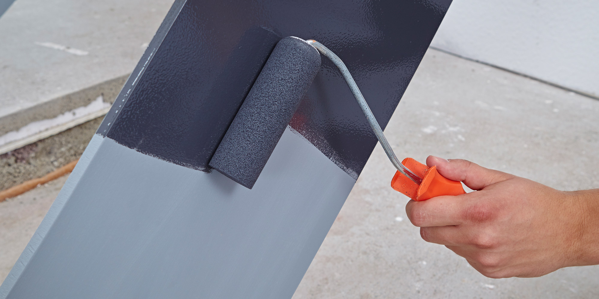 Hand rollt dunkelgrauen Lack auf ein Metallprofil, darunter ist bereits eine hellgraue Schicht aufgetragen