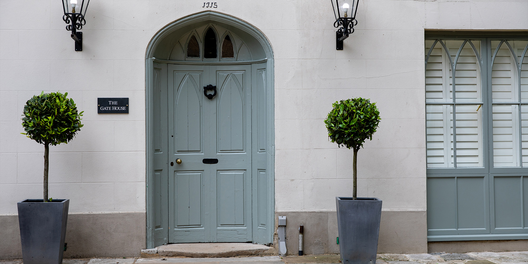 Graue Eingangstür mit Bogenrahmen, zwei Laternen und Kübelpflanzen an heller Fassade