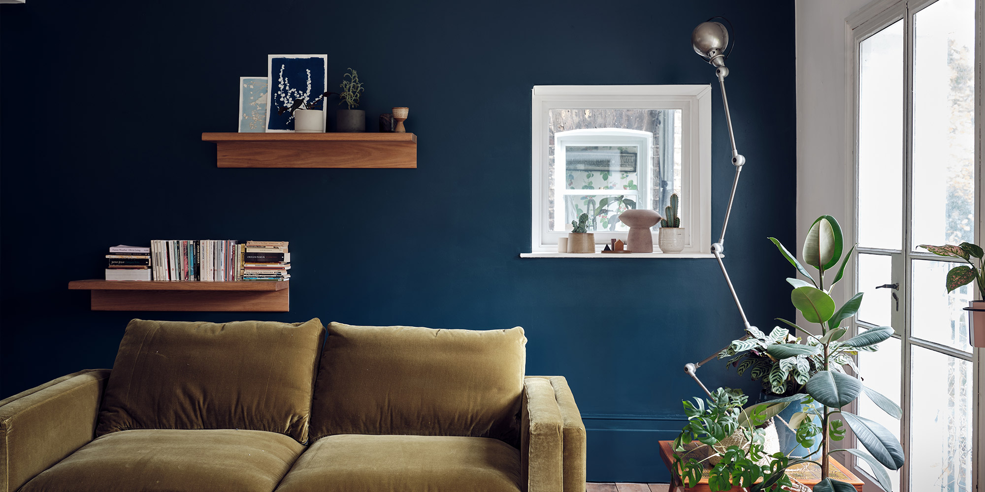 Wohnzimmer mit dunkelblauer Wandfarbe, Sofa, Regalen und Pflanzen am Fenster
