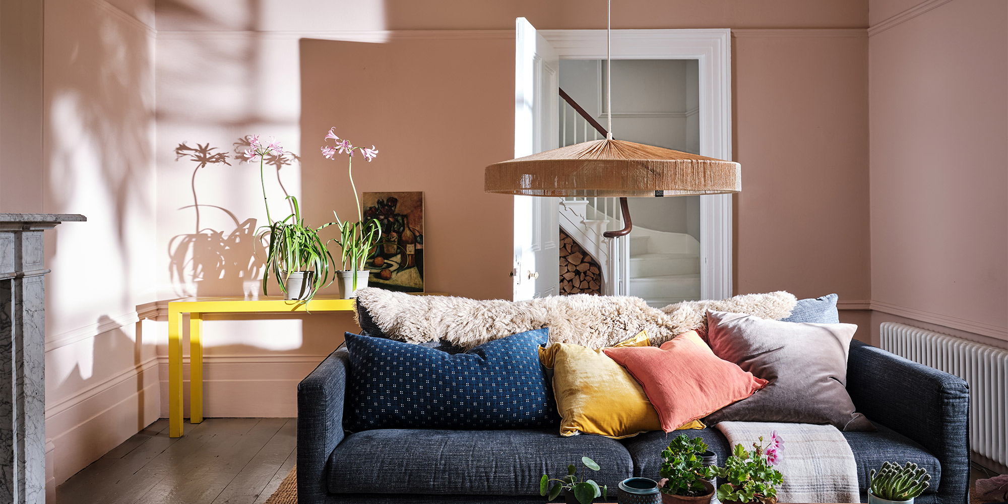 Wohnzimmer mit rosafarbener Wand, blauem Sofa und bunten Kissen unter großer Pendelleuchte