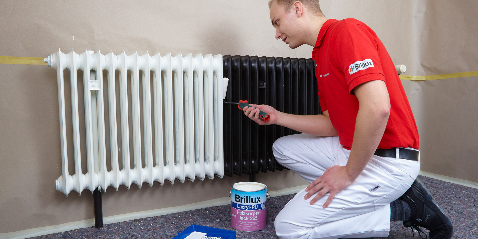 Handwerker lackiert Heizkörper mit kleiner Rolle, daneben Farbdose und Farbwanne