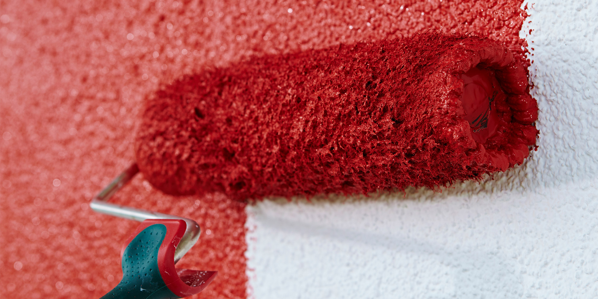 Roter Farbroller streicht eine strukturierte Fassade, daneben bleibt ein weißer Bereich