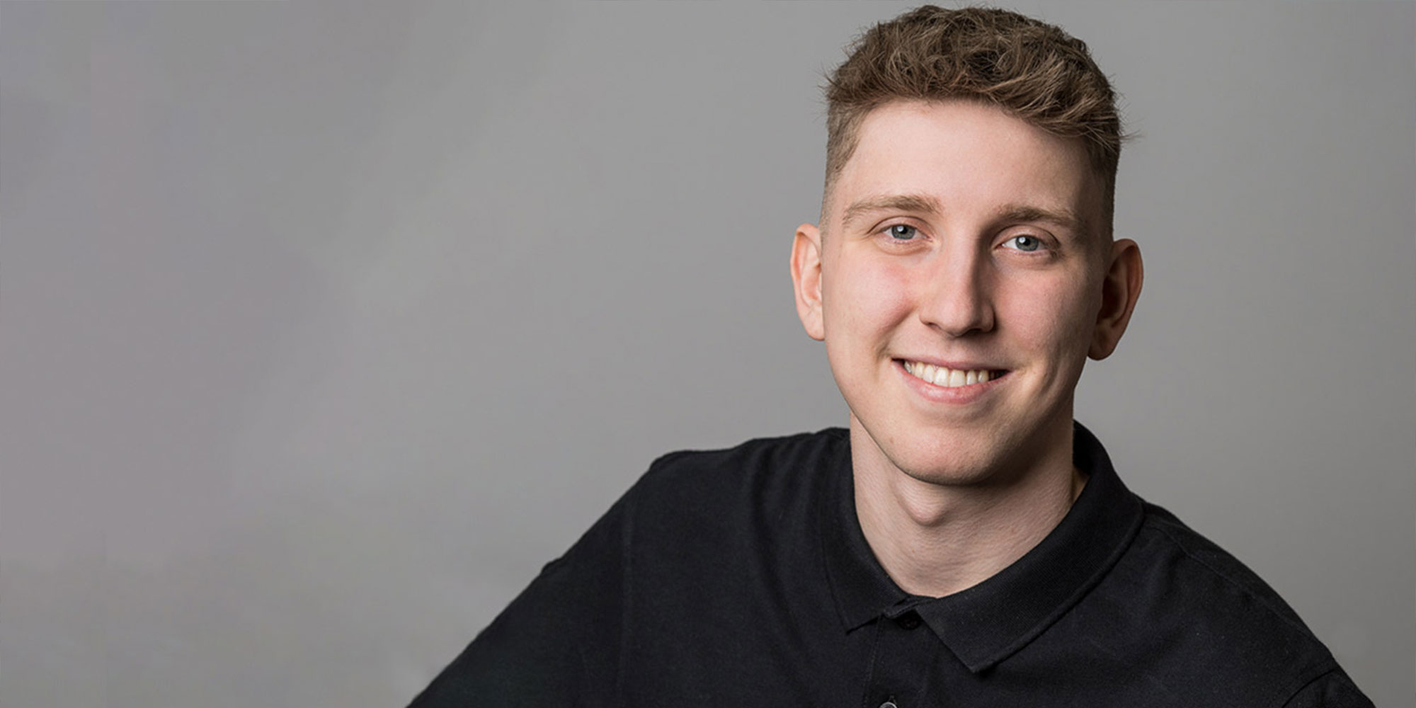 Porträt eines jungen Mannes mit kurzem Haar und schwarzem Hemd, freundlich lächelnd vor neutralem Hintergrund