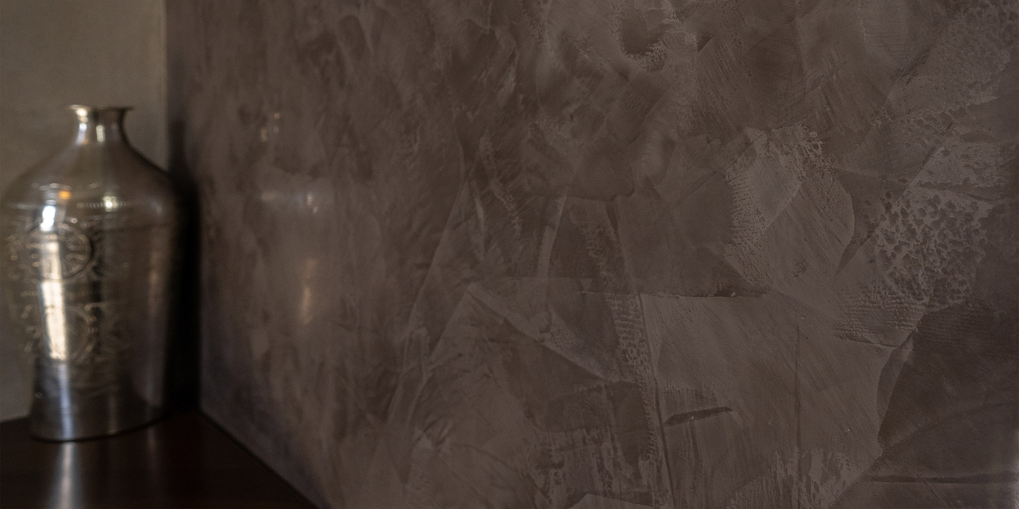 Detailaufnahme einer dunkelbraunen Wand mit strukturierter Frescolori Oberfläche, daneben eine silberne Vase