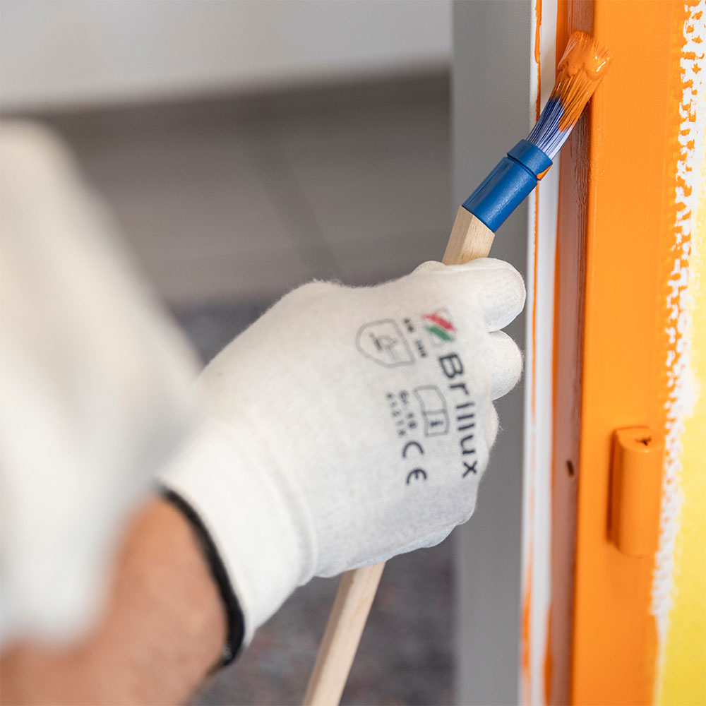 Hand mit Schutzhandschuh streicht orange Lack an eine Fensterrahmenkante, praezise Detailarbeit