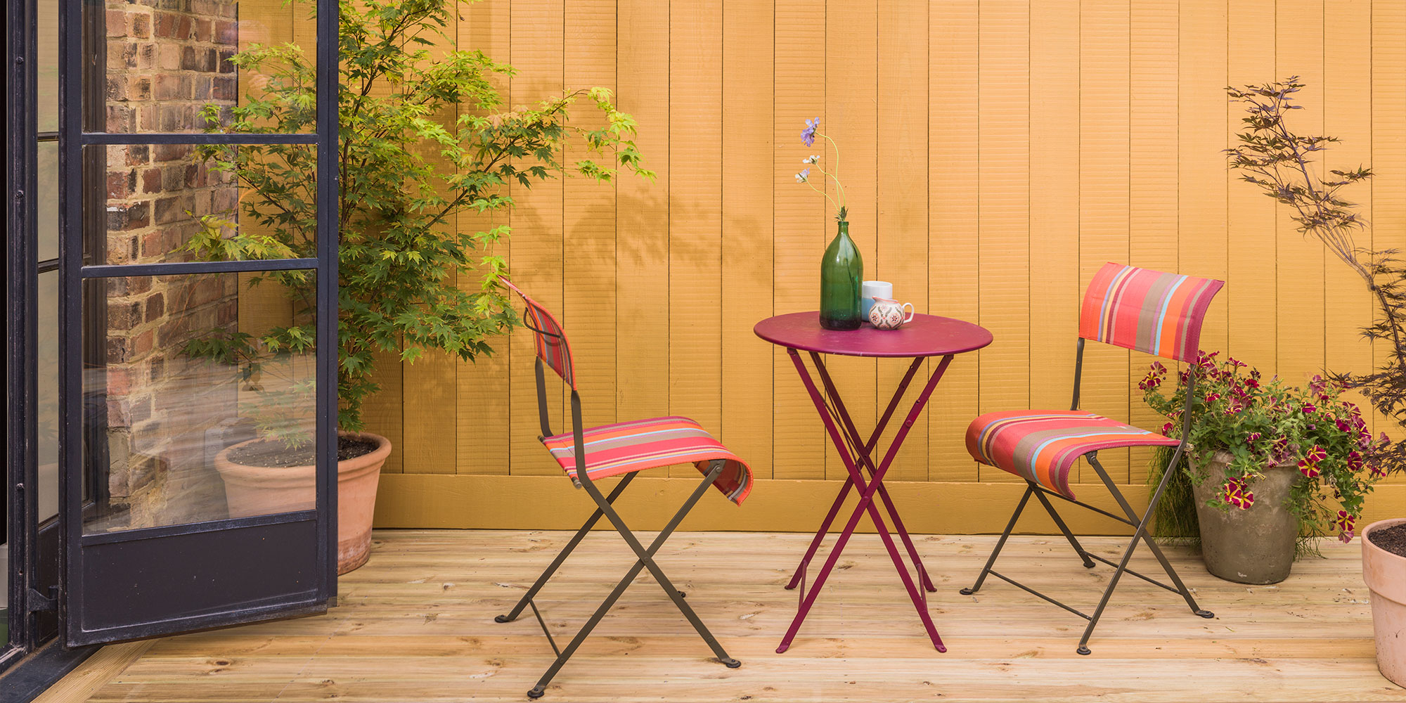 Holzterrasse mit gelb gestrichener Holzwand und Bistroset, Beispiel für Holzschutz und Wetterschutz im Außenbereich.