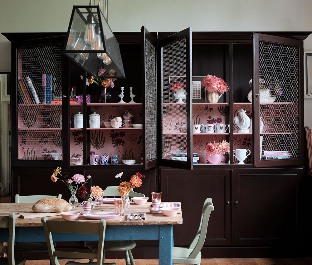Dunkelbraun gestrichener Wohnzimmerschrank mit geöffneten Gittertüren und rosafarbenen Regalböden am Esstisch