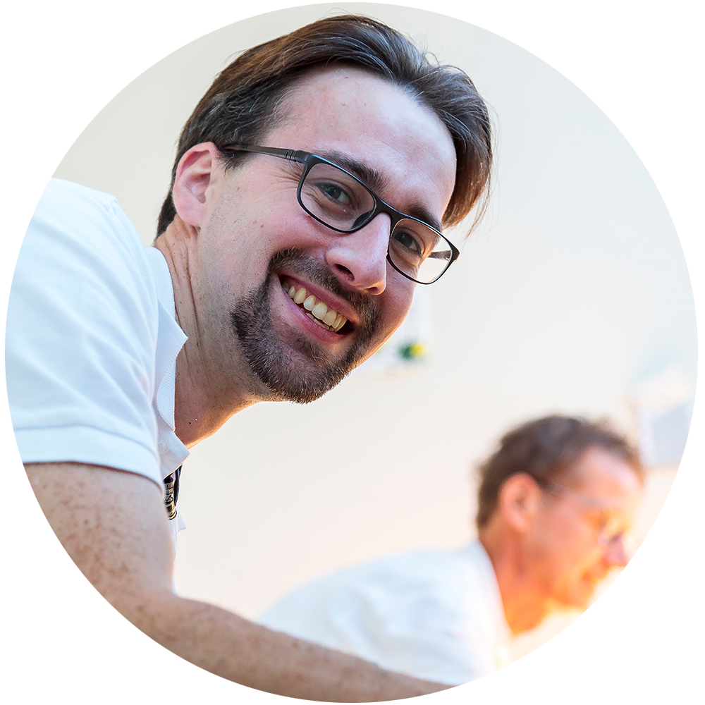 Porträt eines Malers mit Brille und Bart, lächelnd in Arbeitskleidung vor hellem Hintergrund