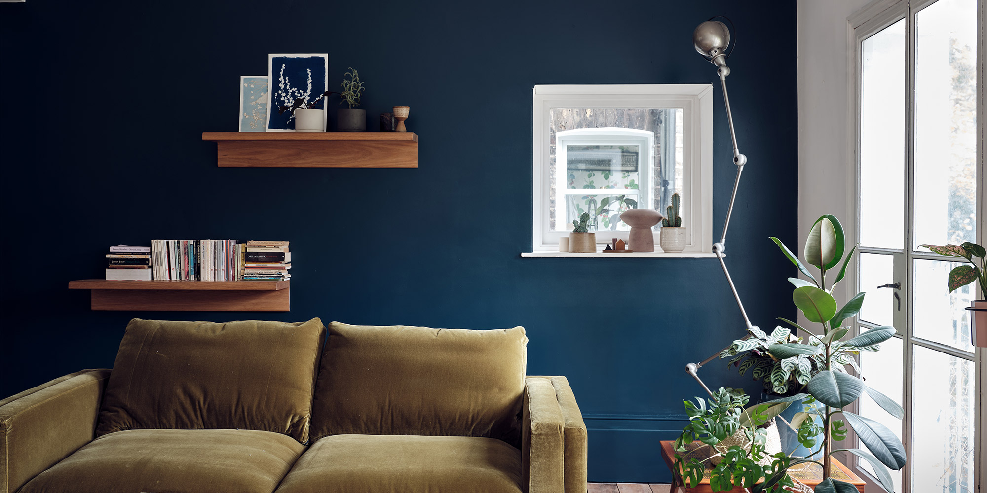 Wohnzimmer mit dunkelblauer Wand, grünem Sofa, Wandregalen und Pflanzen am Fenster