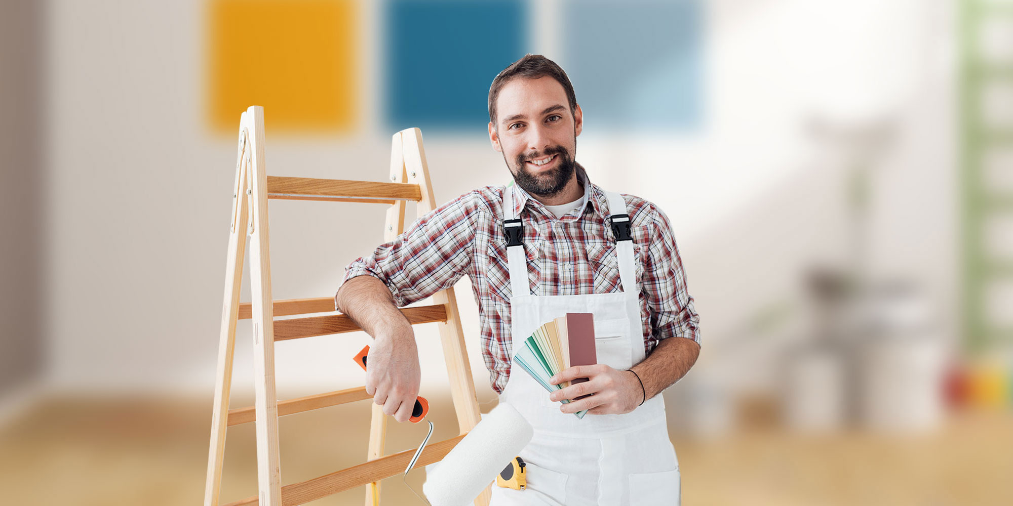 Handwerker mit Bart und kariertem Hemd steht vor Leiter, hält Farbroller und Farbfächer, im Hintergrund ein heller Raum mit Farbmuster an der Wand
