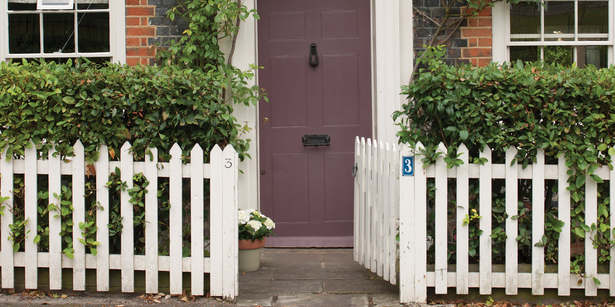 Dunkle Haustür hinter weißem Lattenzaun mit grüner Hecke und Eingangsweg