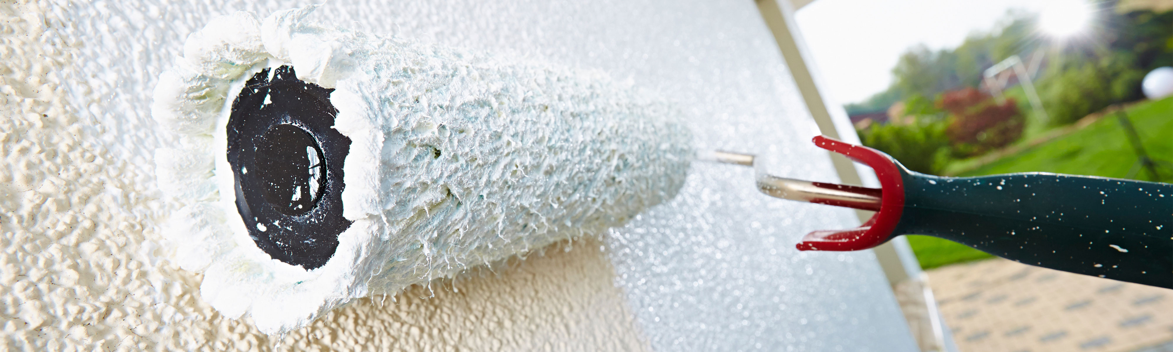 Farbroller trägt weiße Beschichtung auf strukturierten Putz auf, schräge Perspektive im Außenbereich