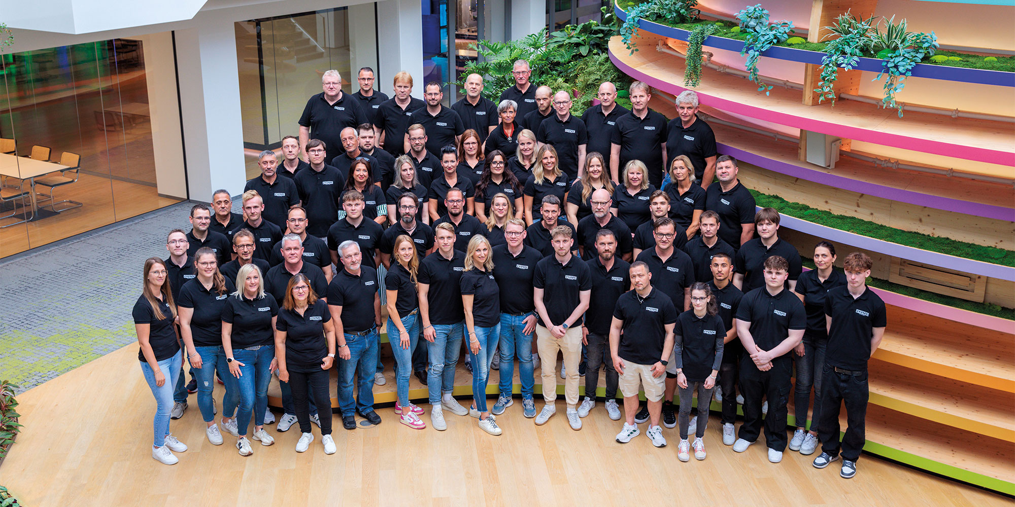 Großes Teamfoto von Mitarbeitenden in schwarzer Kleidung, aufgenommen in einem modernen, hellen Gebäude mit Pflanzen