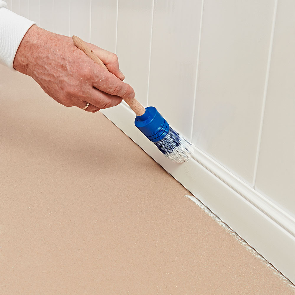 Hand streicht weiße Sockelleiste mit Pinsel am Boden entlang - sauberes Finish beim Bodenanstrich