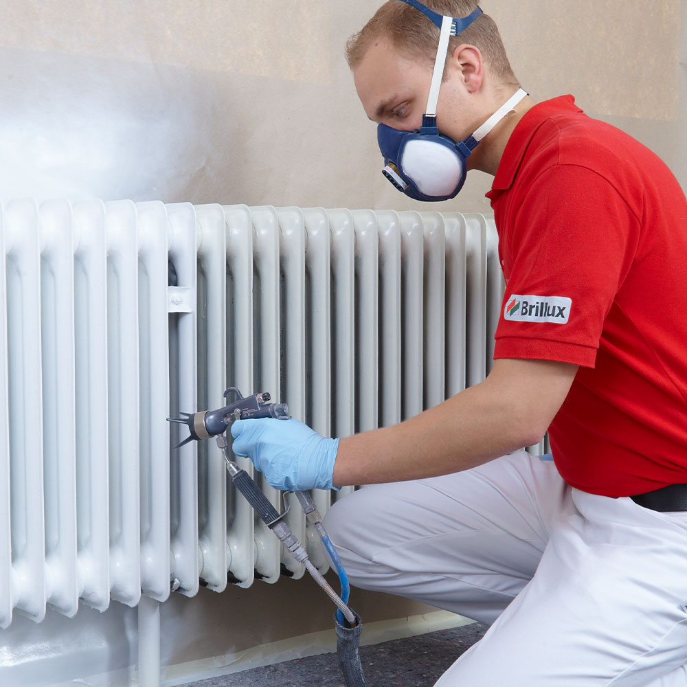 Maler mit Atemschutz sprueht einen Heizkoerper weiß - sicherer Auftrag beim Heizkoerper streichen