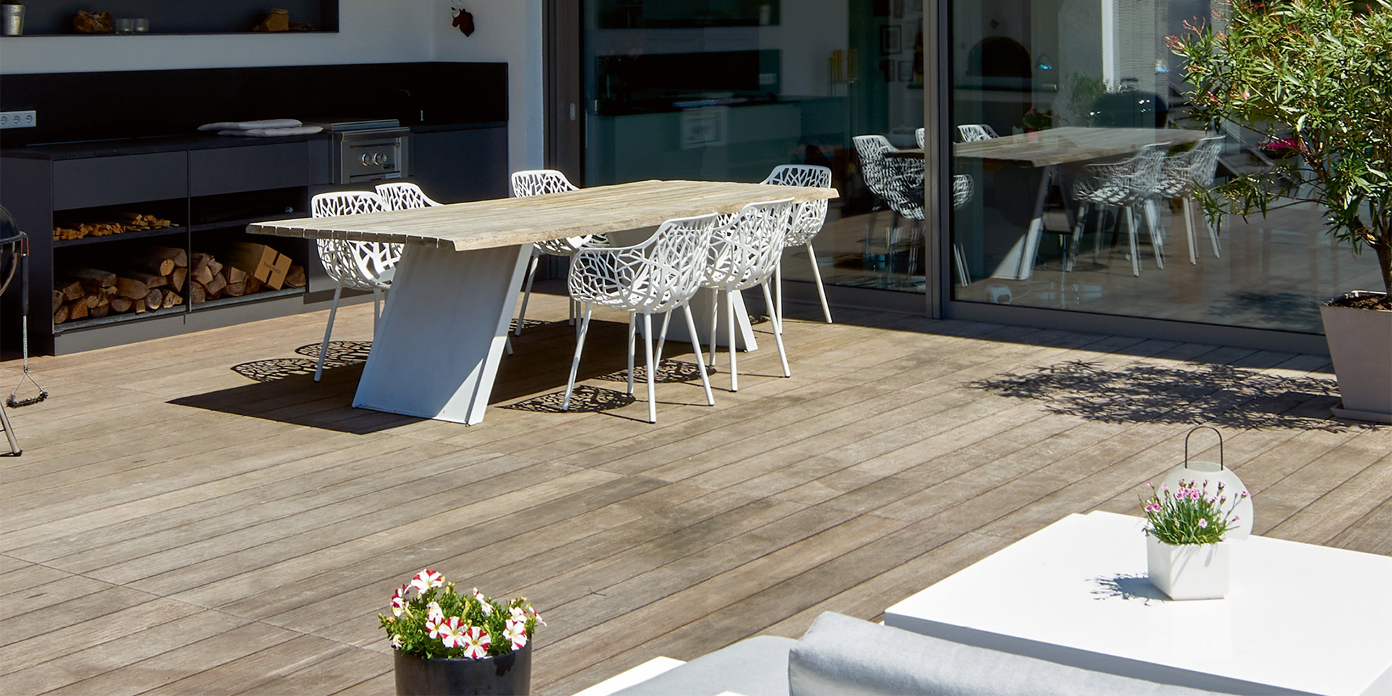 Terrasse mit Outdoor-Küche, Holztisch und weißen Stühlen vor Glasfront