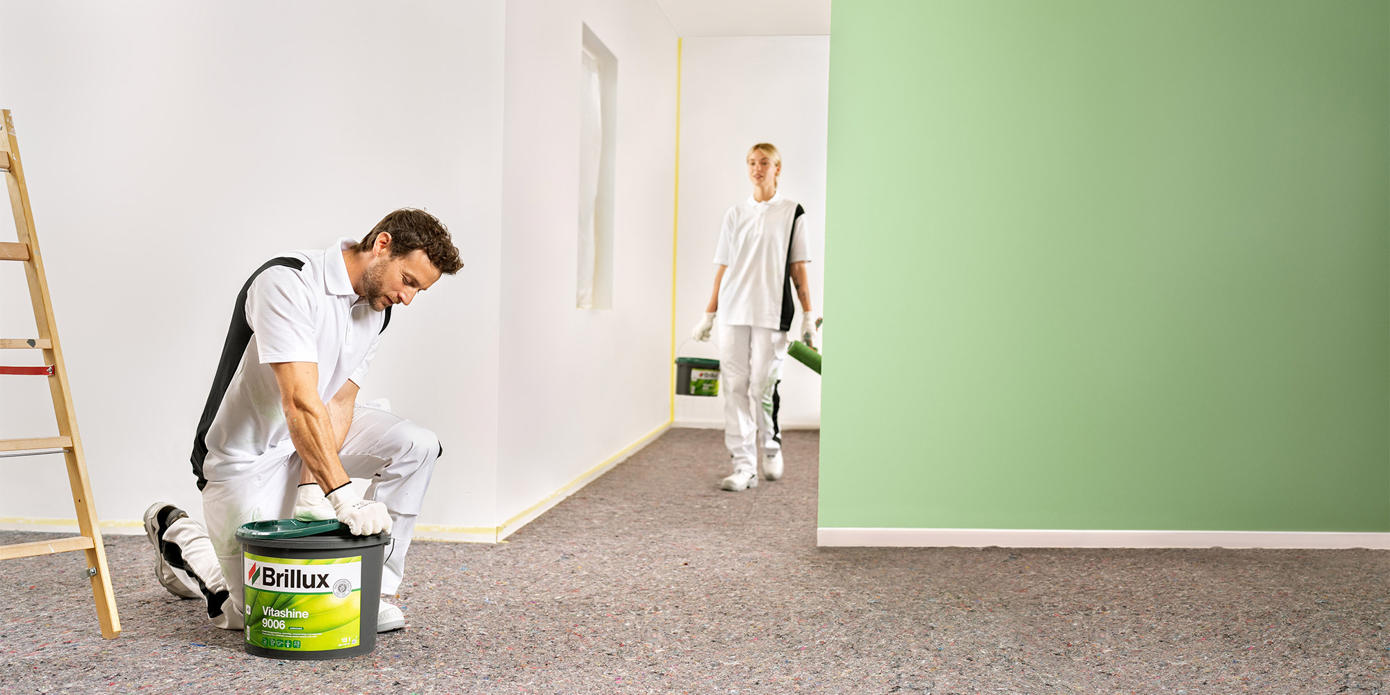 Zwei Maler in einem Raum mit grün gestrichener Wand, Brillux Farbeimer am Boden