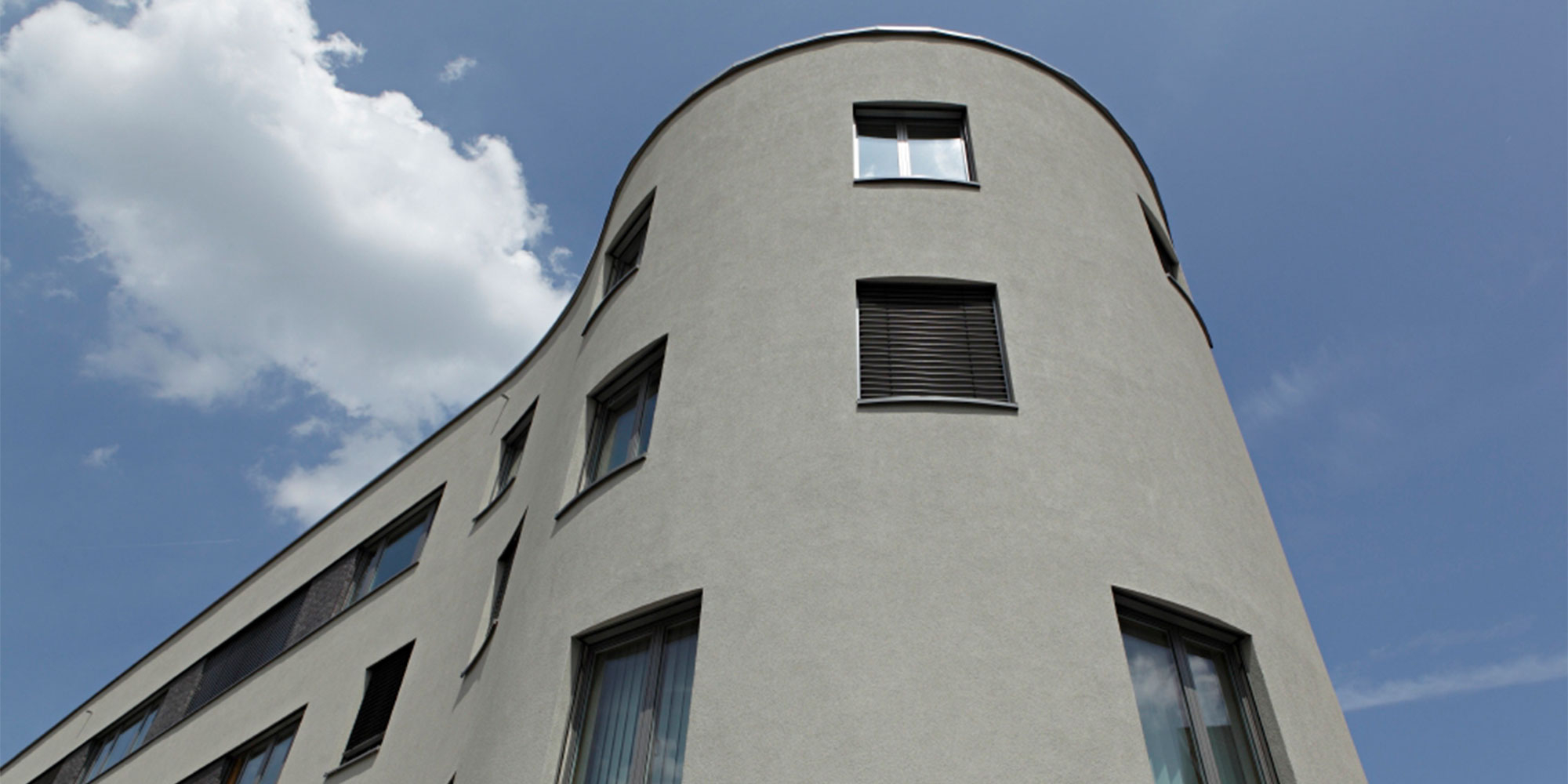 Mehrgeschossiges Gebäude mit abgerundeter Ecke und grauer Putzfassade unter blauem Himmel