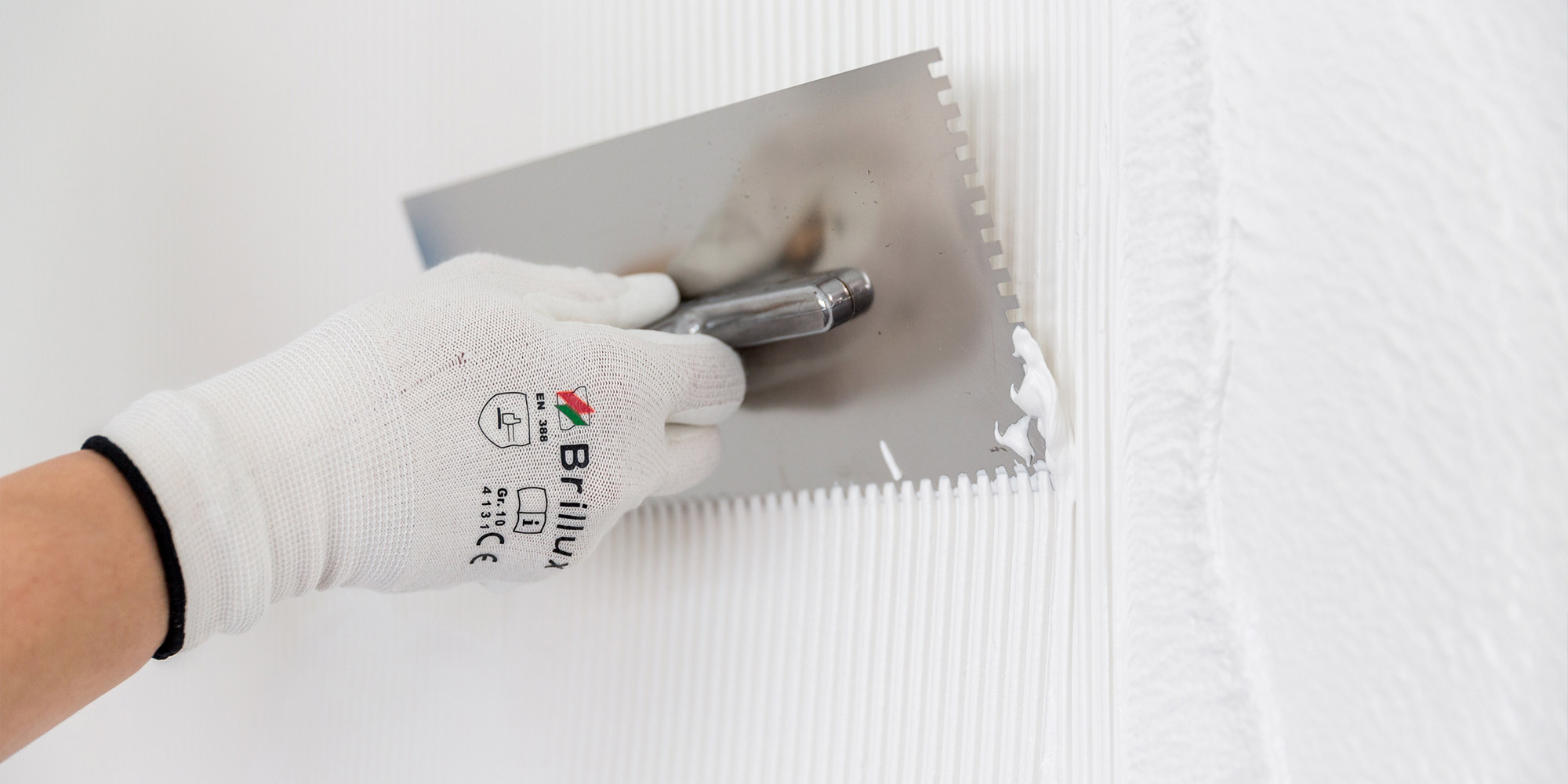 Hand zieht mit Zahnspachtel Kleber auf einer Wandflaeche auf