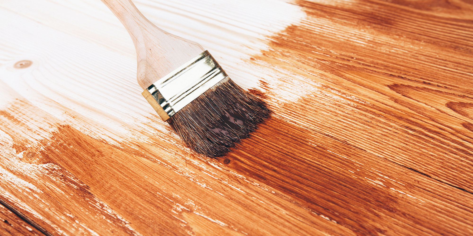 Breiter Pinsel streicht braune Lasur ueber ein Holzbrett, links bleibt ein heller Bereich unbeschichtet