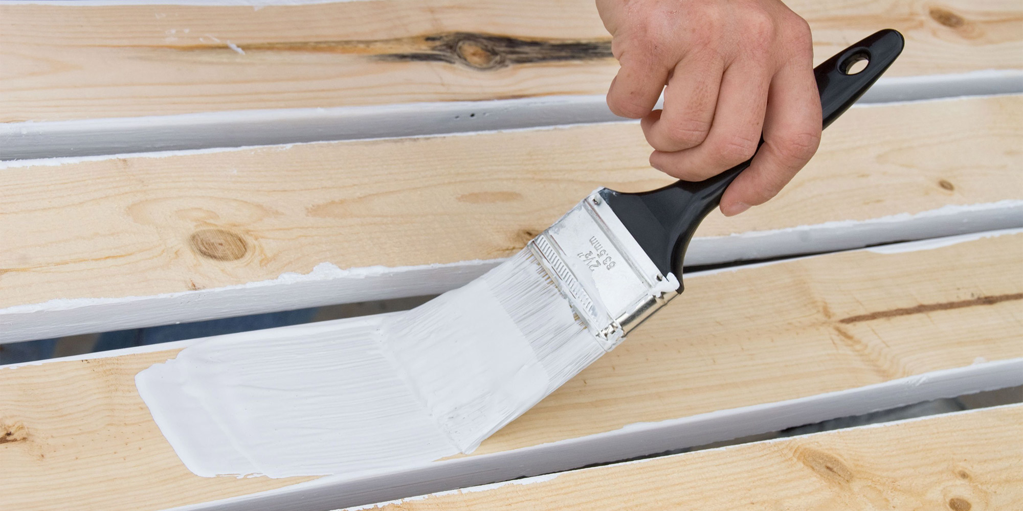 Hand trägt mit einem Pinsel weiße Farbe auf helle Holzbretter auf, gleichmäßiger Farbauftrag sichtbar