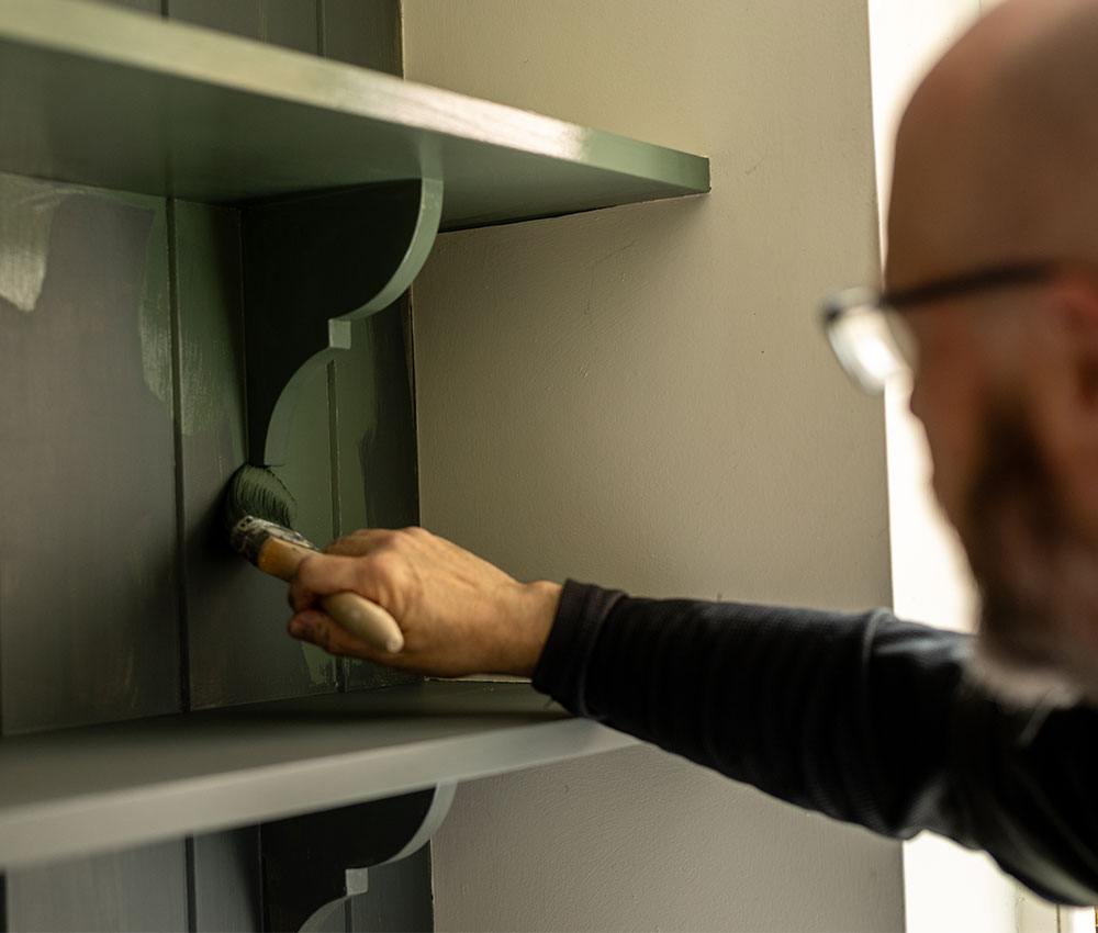 Hand streicht ein Holzregal mit Pinsel in Dunkelgrün, Detailaufnahme beim Möbel streichen innen.