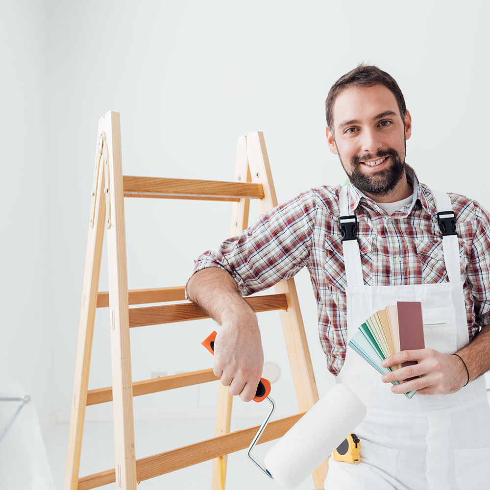 Handwerker in weißer Latzhose steht vor einer Holzleiter, hält einen Farbroller und einen Farbfächer, heller Raum im Hintergrund