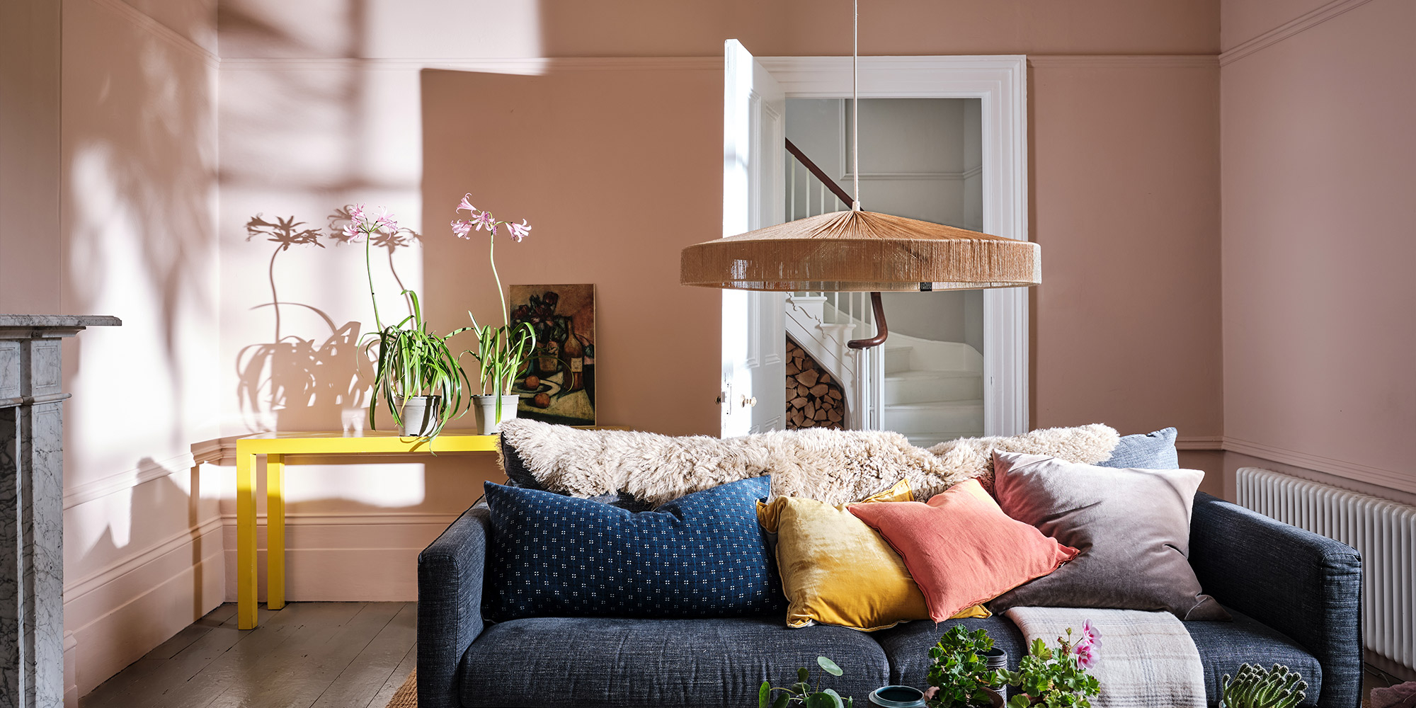 Wohnbereich mit zartrosa Wandfarbe, dunklem Sofa und farbigen Kissen im Tageslicht